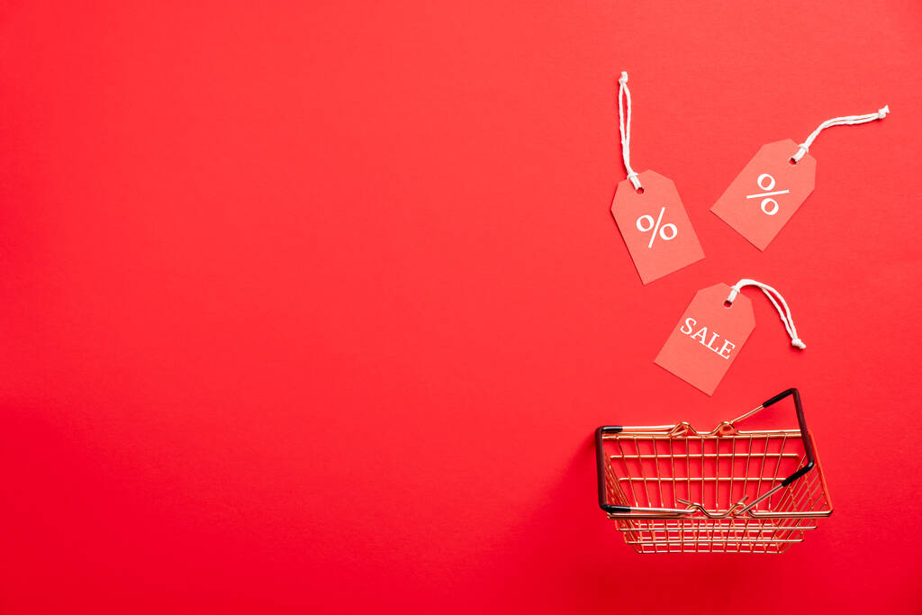 Shopping basket and tags on red background. Black Friday Sale concept. Flat lay, top view. - Photo, Image