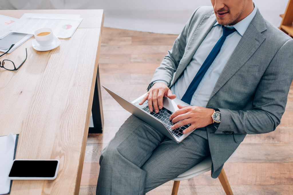 Обрезанный вид бизнесмена с помощью ноутбука рядом со смартфоном и кофе на офисном столе  - Фото, изображение