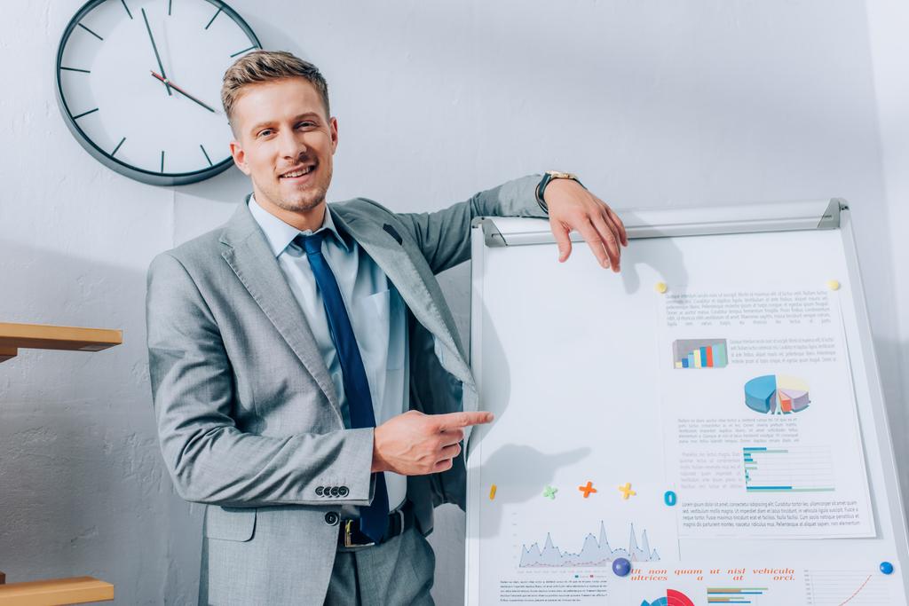 Smiling businessman pointing at papers with graphs on flipchart in office  - Photo, Image