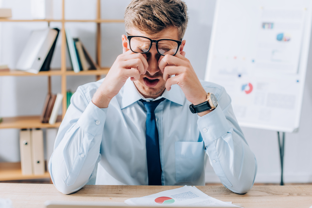 Бизнесмен в очках теребит усталые глаза возле бумаг на столе - Фото, изображение