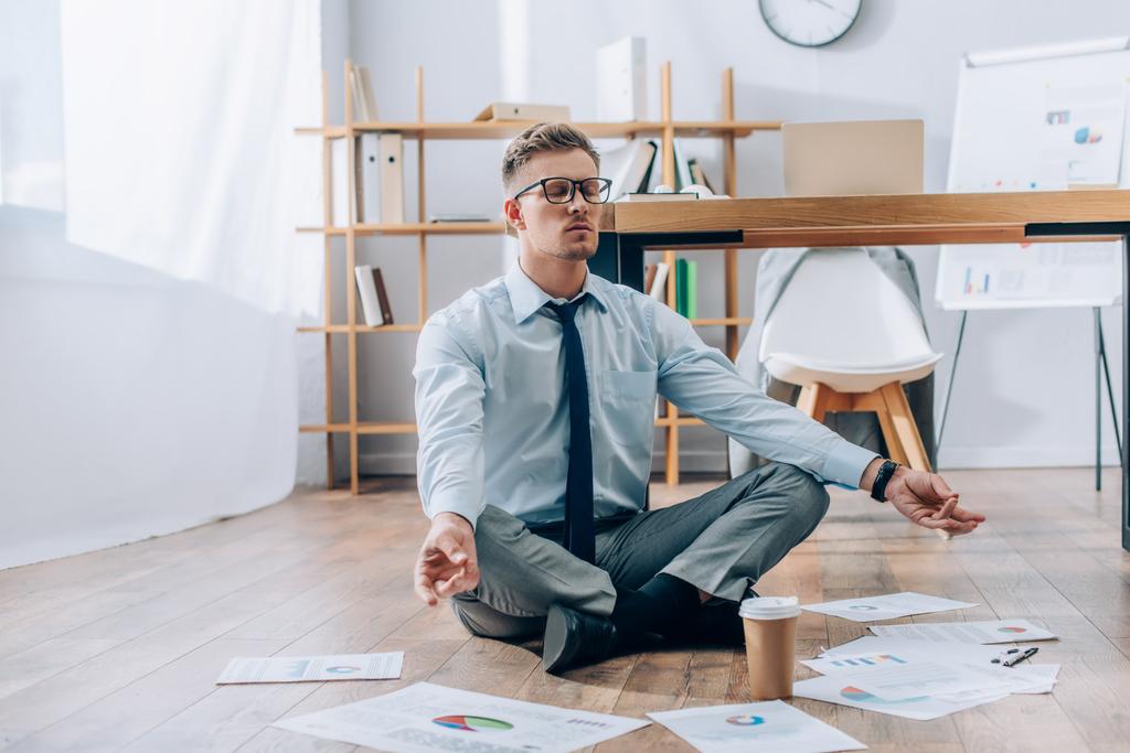 Jovem empresário meditando perto de papéis e café para ir ao chão no escritório  - Foto, Imagem