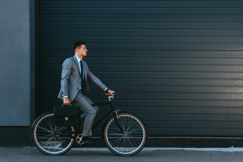 Молодой бизнесмен с портфелем на велосипеде возле фасада здания  - Фото, изображение