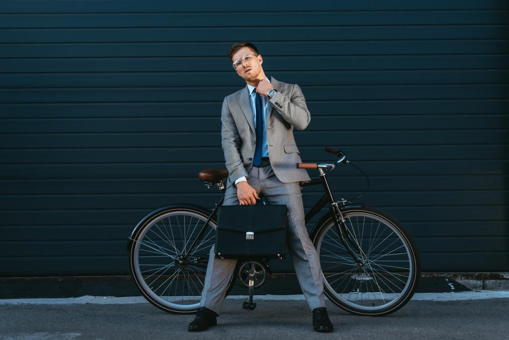 Jeune homme d'affaires avec mallette regardant la caméra près du vélo à l'extérieur  - Photo, image