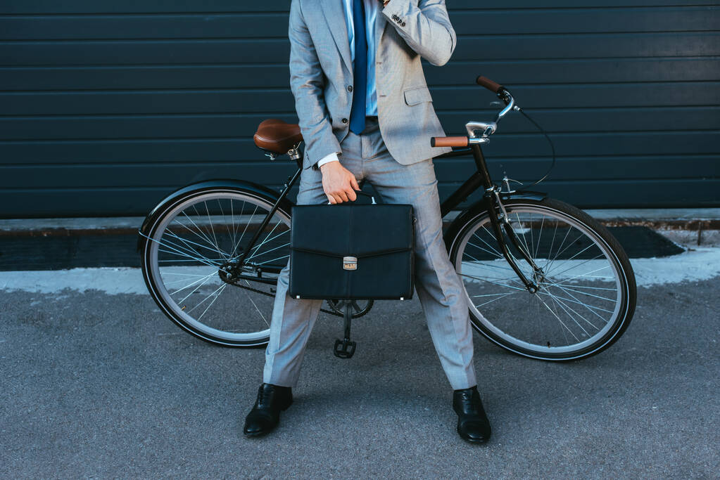 Vista recortada del hombre de negocios sosteniendo el maletín mientras está de pie cerca de la bicicleta y el edificio al aire libre  - Foto, Imagen