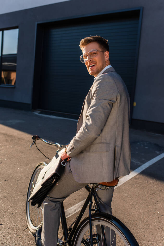 Νεαρός επιχειρηματίας χαμογελά στην κάμερα, ενώ κρατώντας χαρτοφύλακα σε ποδήλατο σε εξωτερικούς χώρους  - Φωτογραφία, εικόνα