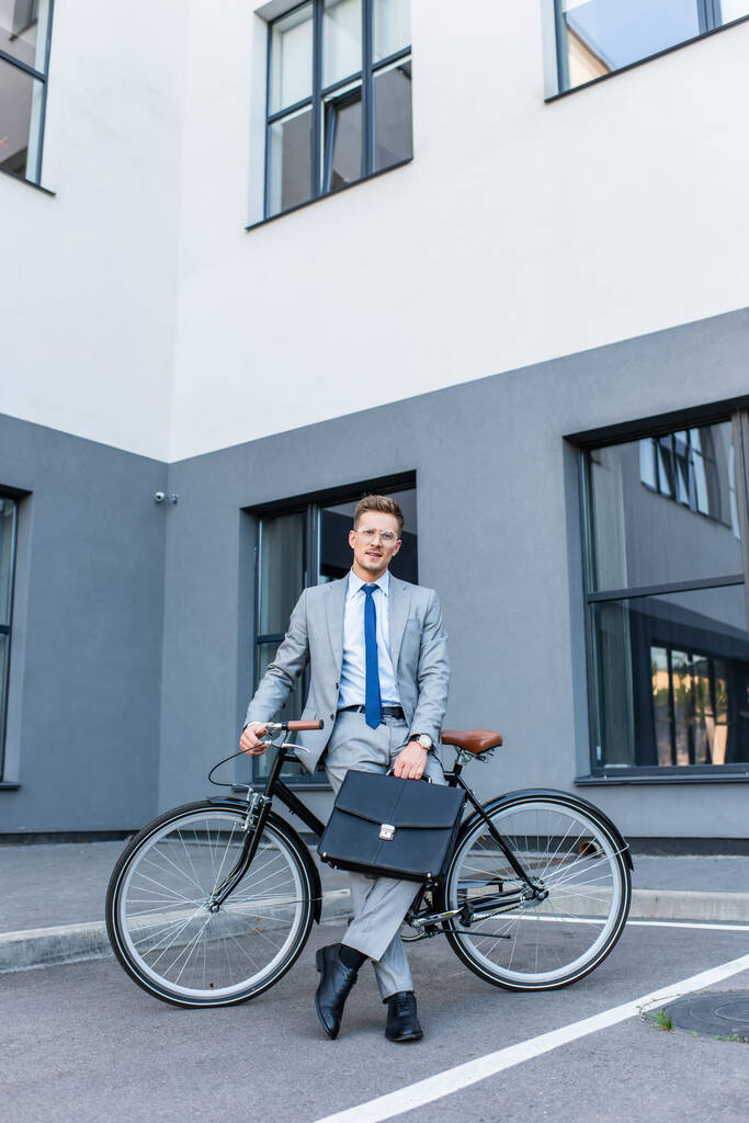 眼鏡やスーツのビジネスマンは、自転車や建物の近くにブリーフケースを保持  - 写真・画像