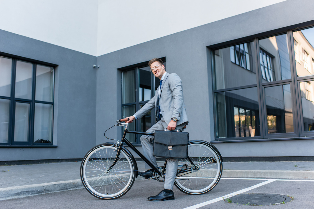 Üzletember mosolyog a kamerába, miközben aktatáskát tart és kerékpározik a szabadban  - Fotó, kép