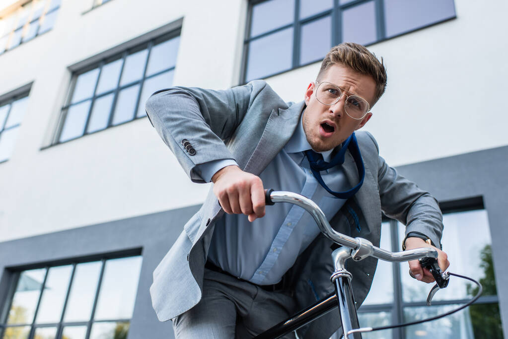 Заплутаний бізнесмен дивиться на камеру під час їзди на велосипеді біля будівлі
  - Фото, зображення