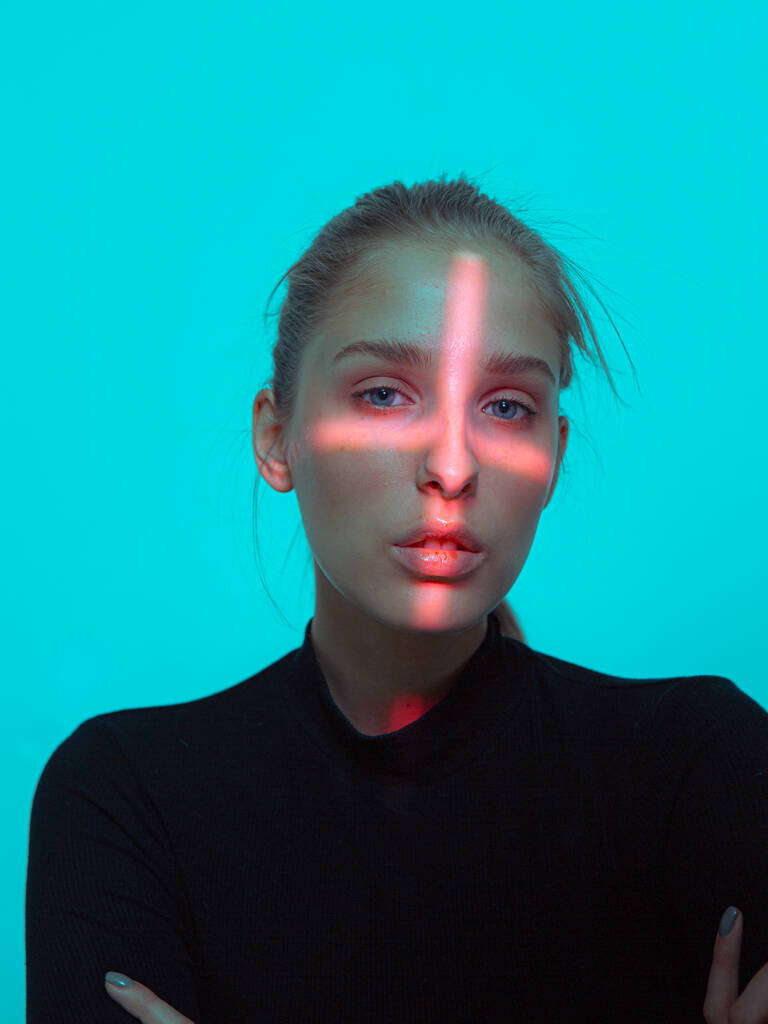 Портрет молодой красивой девушки с крестообразным красным светом на ее лице на синем фоне студии - Фото, изображение
