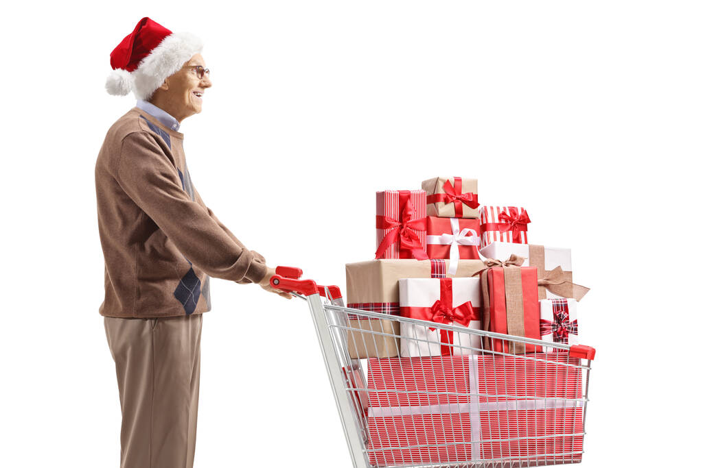 サンタクラスの帽子をかぶった高齢者やクリスマスプレゼント付きのショッピングカートは、白い背景に隔離されています - 写真・画像