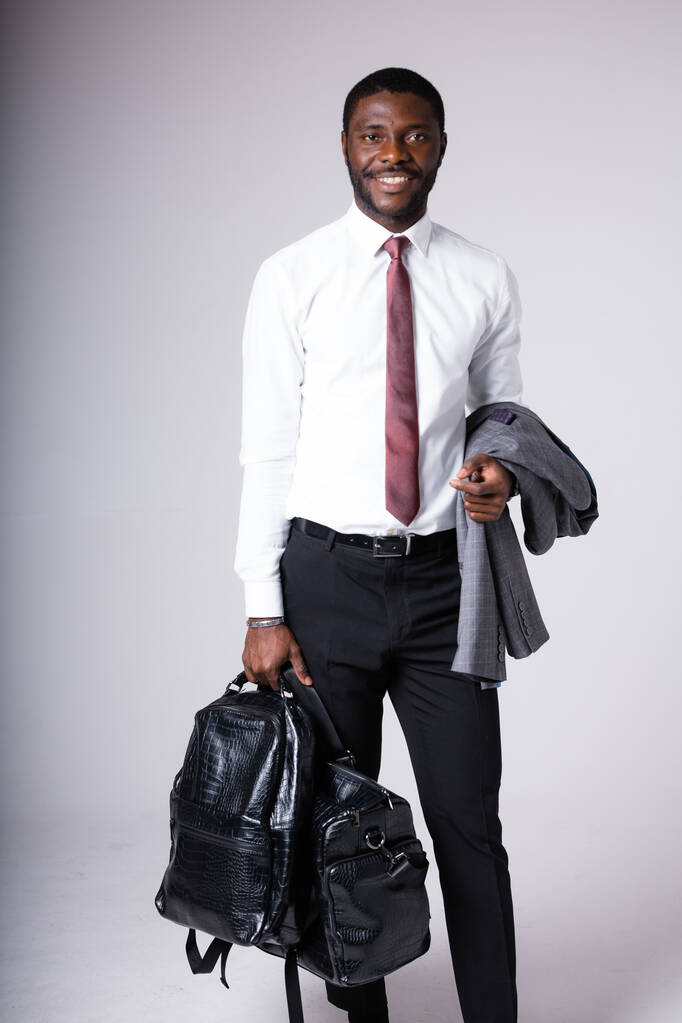 Афроамериканець у класичній білій сорочці і чорних штанях стоїть на сірому фоні, тримаючи чорну сумку для подорожей і сіру куртку. - Фото, зображення