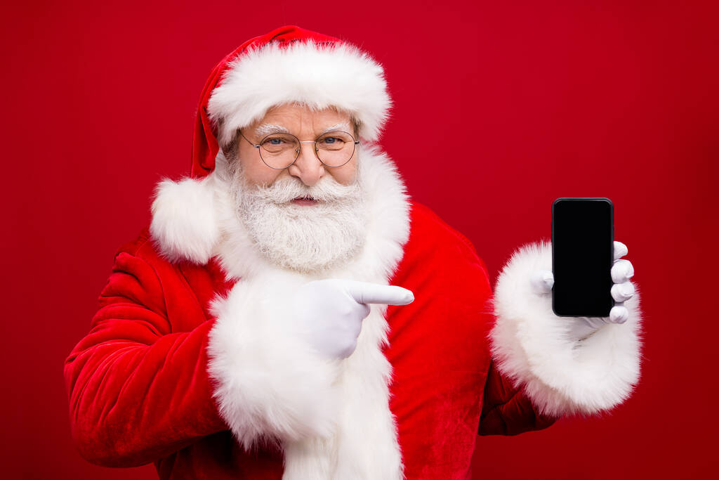 Zdjęcie emeryta dziadek szary broda trzymać telefon puste miejsce bezpośredni ekran palca sugerują sprzedaż produkt x-mas kostium białe rękawiczki płaszcz okulary nakrycia głowy odizolowany czerwony kolor tło - Zdjęcie, obraz