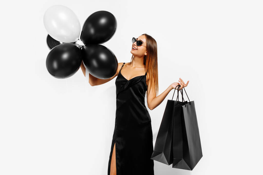 Kara Cuma indirimi. Beyaz arka planda balonları ve alışveriş çantaları olan siyah şık elbiseli genç bir kadın. - Fotoğraf, Görsel