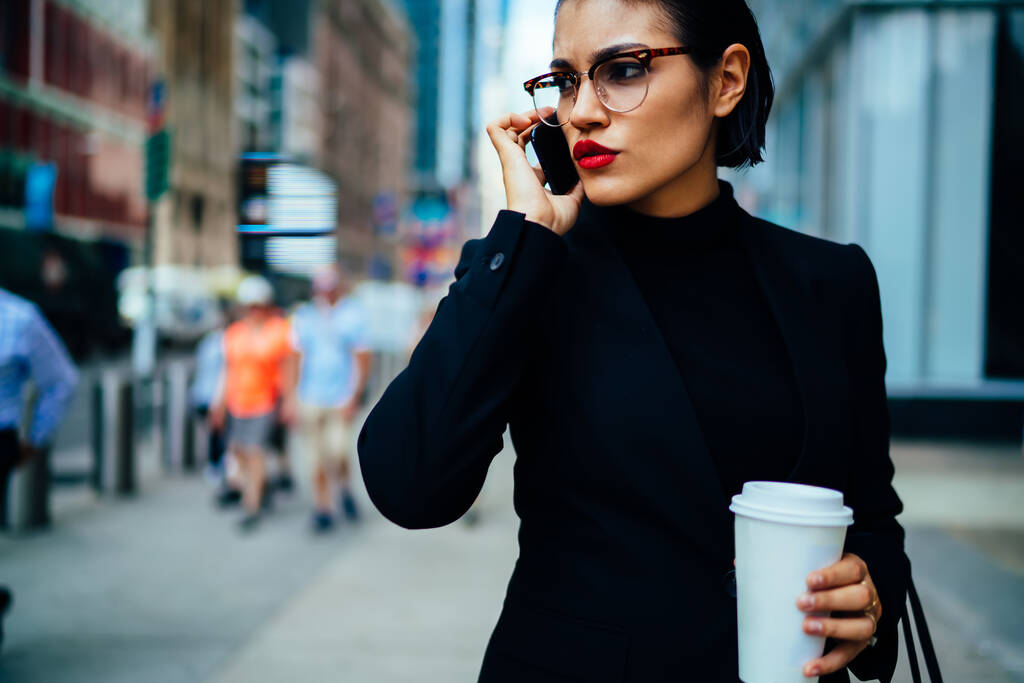 Recadrer sérieux confiant entrepreneur féminin habillé en costume élégant parler sur téléphone portable tout en se tenant debout avec une tasse de café pour aller dans la rue - Photo, image
