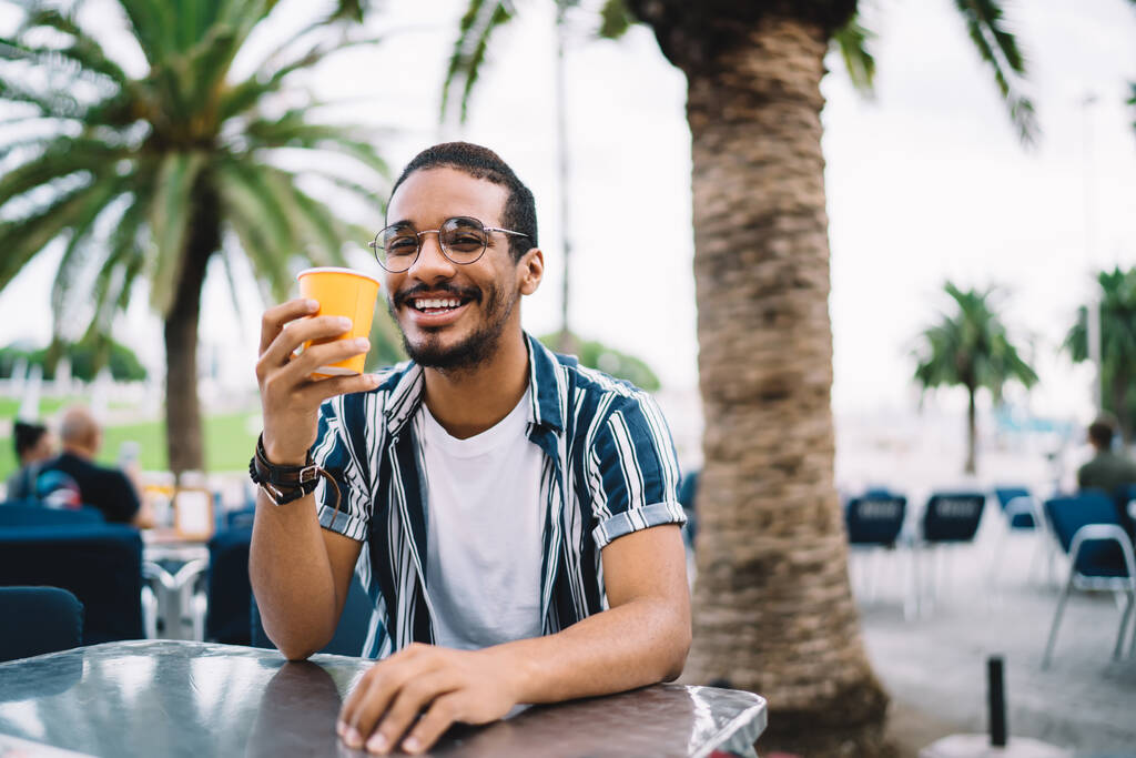 Puoli pituus muotokuva iloinen afrikkalainen amerikkalainen hipster kaveri rento kulumista juominen juoma kaupungin uudelleen loma lomat, onnellinen teini mies silmälasit visio korjaus tilalla cup - Valokuva, kuva