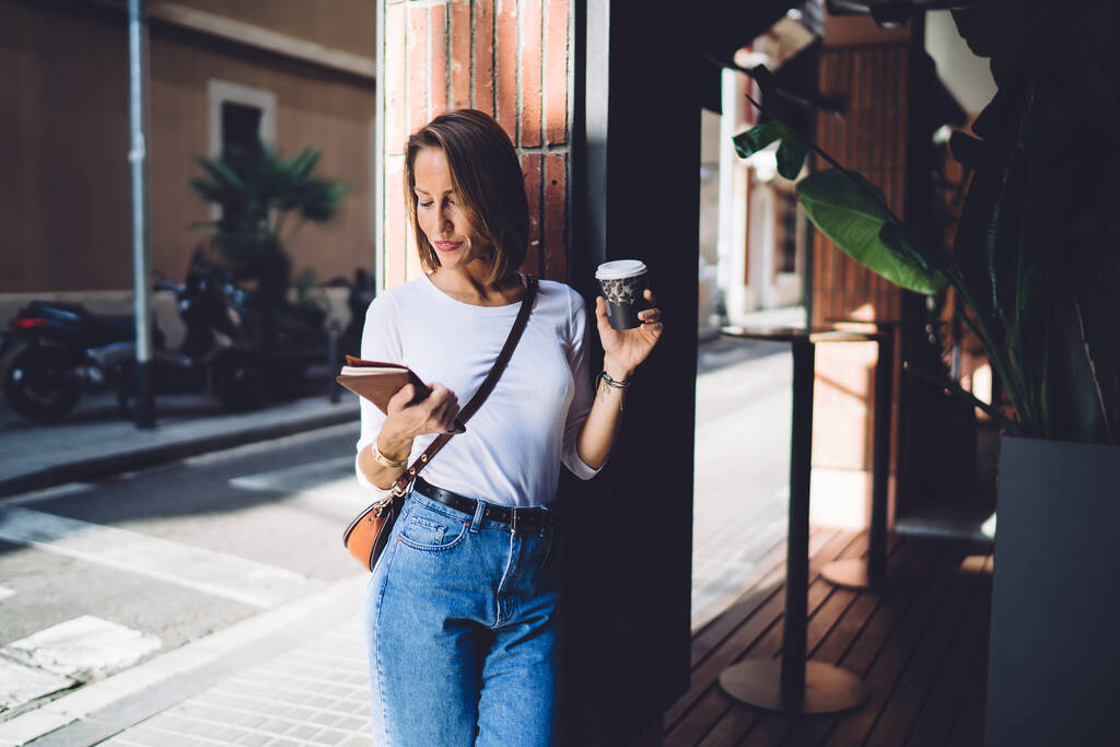Elegante hembra adulta con ropa casual sonriendo y bebiendo café caliente mientras está de pie en la calle con libro en la mano cerca de la cafetería moderna - Foto, Imagen