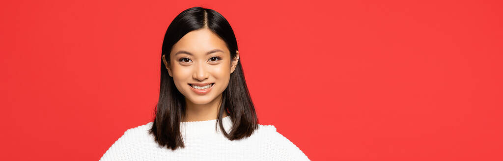 glücklich und junge asiatische Frau im Pullover schaut in die Kamera isoliert auf rotem Banner - Foto, Bild