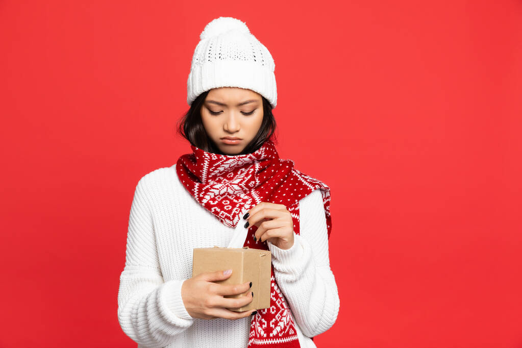 triste asiatique femme dans chapeau et écharpe tenant boîte de tissu isolé sur rouge  - Photo, image