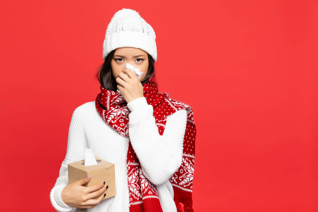 enfermo asiático mujer en sombrero y bufanda celebración servilleta y tejido caja mientras estornudos aislado en rojo  - Foto, imagen