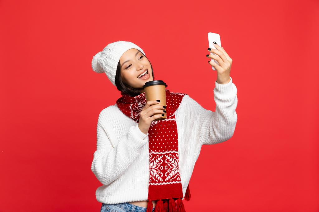 aufgeregte Asiatin mit Hut und Schal hält Einwegbecher in der Hand und macht ein Selfie auf rotem Grund  - Foto, Bild