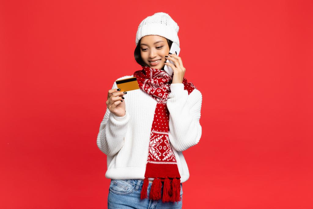 sonriente asiático mujer en sombrero y bufanda celebración smartphone y mirando a la tarjeta de crédito aislado en rojo - Foto, Imagen