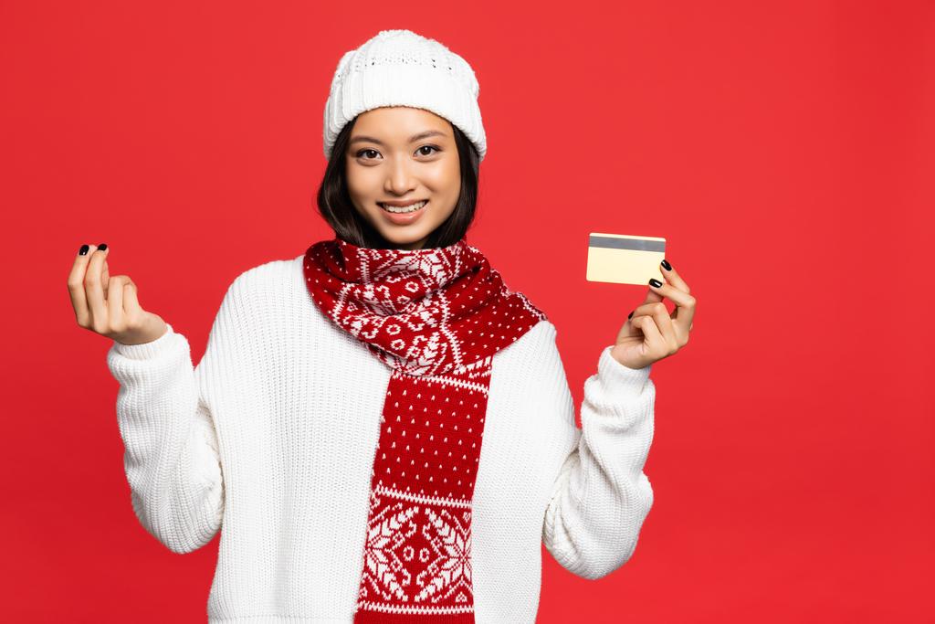 улыбающаяся азиатка в шляпе и шарфе, держащая кредитку и показывающая жест денег, изолированный на красном - Фото, изображение