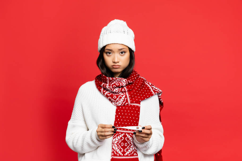 upset and sick asian woman in hat and scarf holding electronic thermometer isolated on red  - Photo, Image