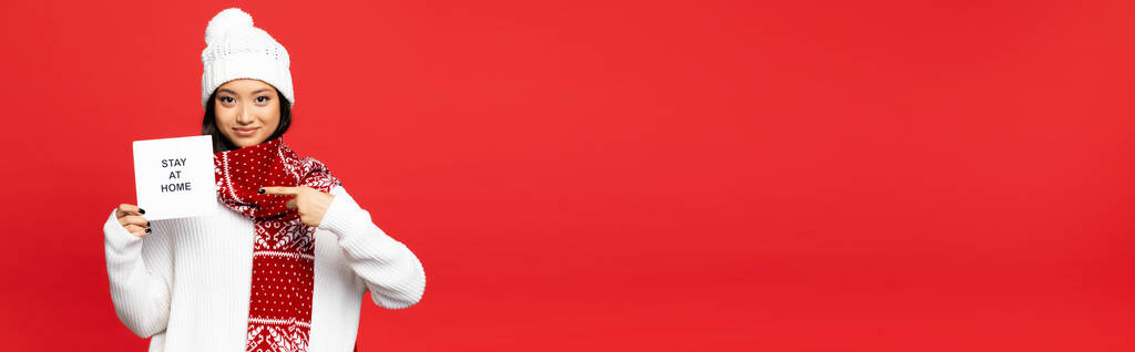 giovane donna asiatica in cappello bianco e sciarpa punta con il dito a carta con soggiorno a casa lettering isolato su rosso, striscione - Foto, immagini