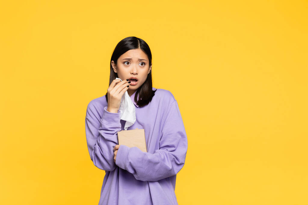 conmocionada mujer asiática sosteniendo caja de tejido y mirando hacia otro lado aislado en amarillo  - Foto, Imagen