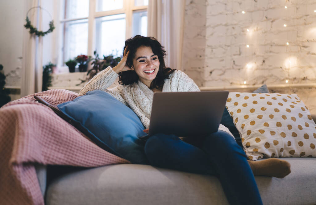 Optimistische vrouw in warme trui en jeans zittend op een comfortabele bank tussen kussens en lachend tijdens het gebruik van laptop in de kersttijd - Foto, afbeelding