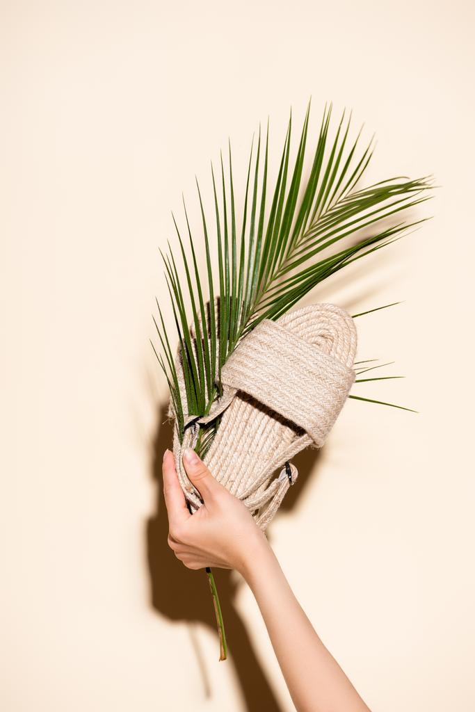 bijgesneden weergave van vrouw met zomer gevlochten sandalen en palmblad op beige achtergrond - Foto, afbeelding