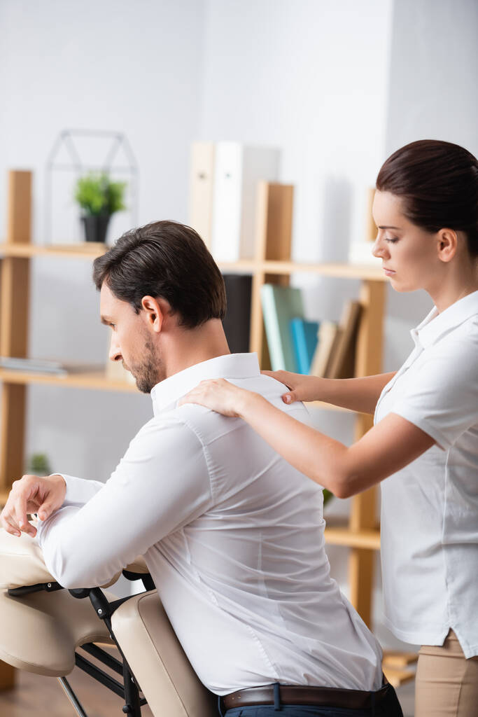 Massaggiatrice massaggiatrice massaggiatrice spalle di uomo d'affari seduto su sedia da massaggio in ufficio su sfondo sfocato - Foto, immagini