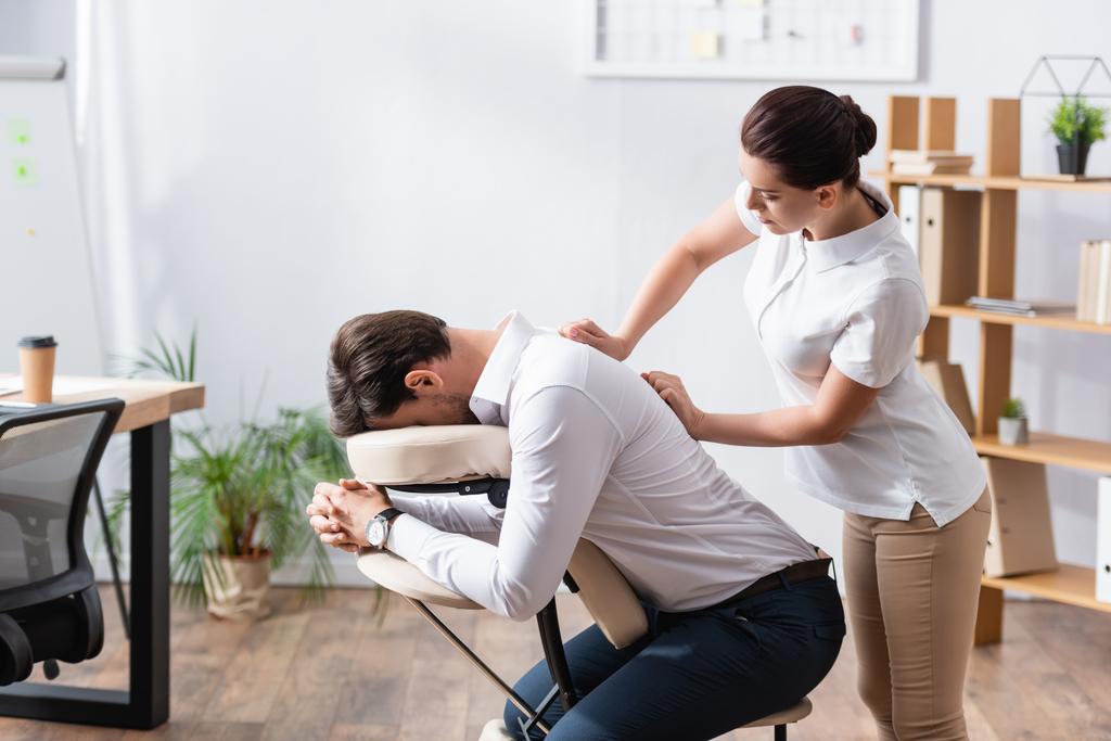 Vista laterale della massaggiatrice massaggiando la schiena dell'uomo d'affari seduto sulla sedia da massaggio in ufficio - Foto, immagini