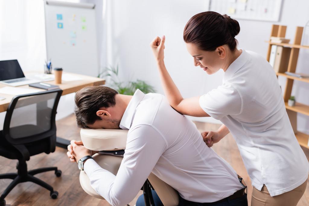 Sorridente massaggiatrice facendo massaggio alla schiena con gomito per uomo d'affari in ufficio su sfondo sfocato - Foto, immagini