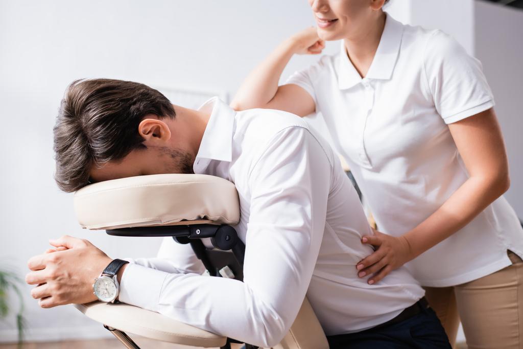 Sorridente massaggiatrice massaggio posteriore di uomo d'affari con gomito in ufficio su sfondo sfocato - Foto, immagini