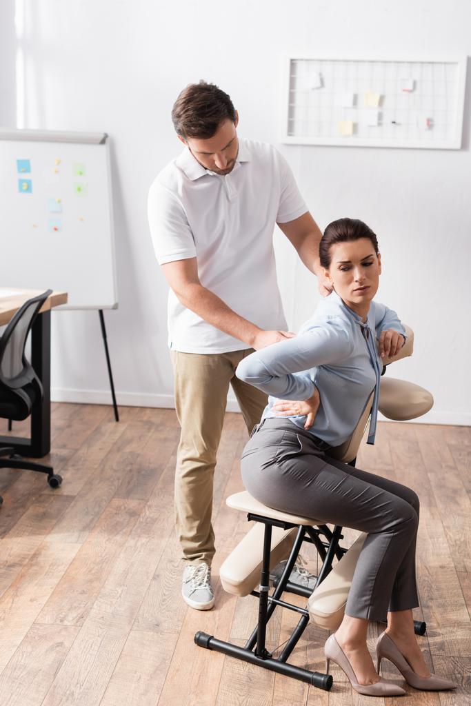 Массер масажує болючу спину жінки з руками на стегні, сидячи на масажному стільці в офісі - Фото, зображення
