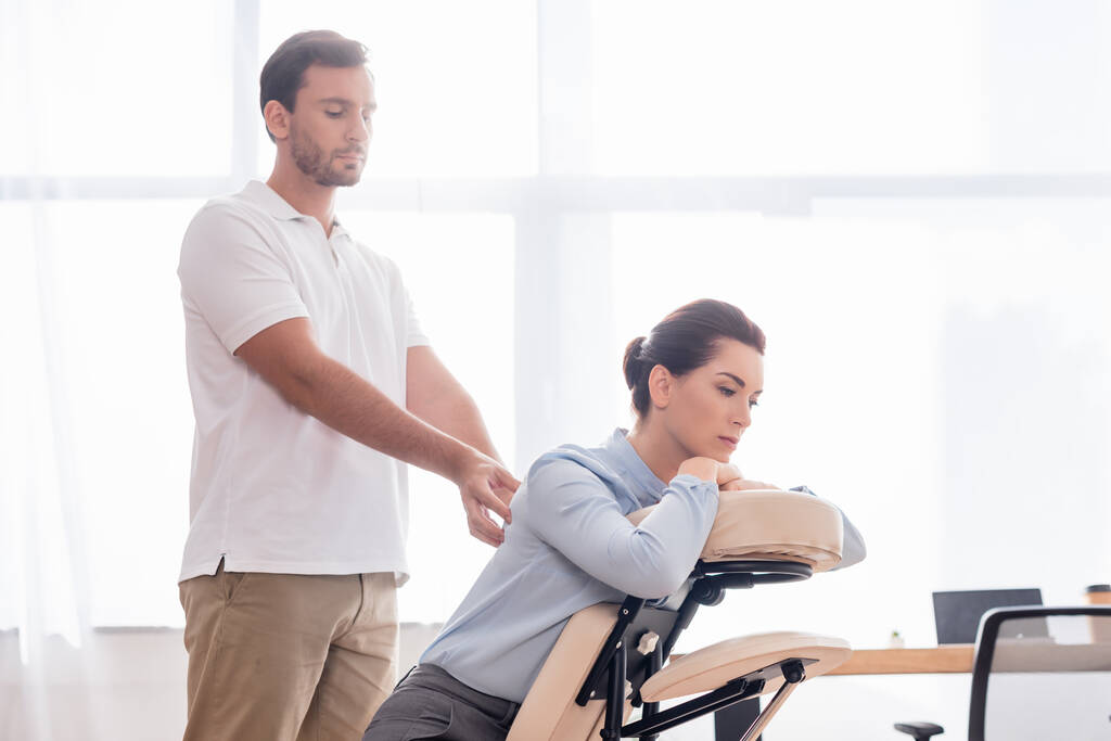 Focused masseur massaging back of brunette of businesswoman sitting on massage chair on blurred background - Photo, Image