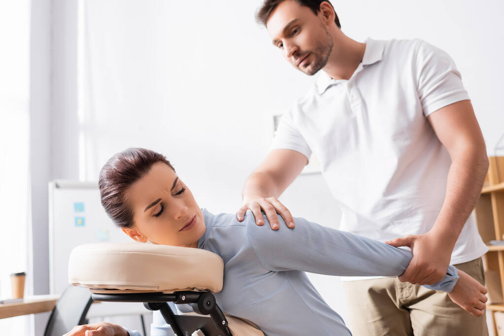 Massage therapist massaging hurting arm of female client sitting on massage chair with blurred office on background - Photo, Image