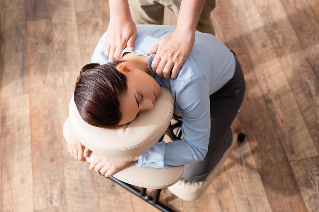 Visão aérea do massagista massageando ombros de empresária morena com olhos fechados, sentada na cadeira de massagem - Foto, Imagem