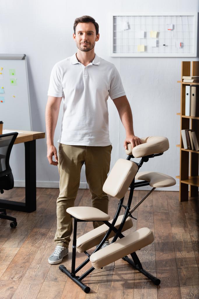 Vista frontale del massaggiatore sorridente guardando la fotocamera, mentre in piedi vicino alla sedia di massaggio in ufficio - Foto, immagini