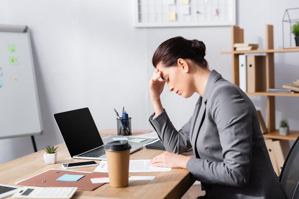 Empresária cansada com a mão na testa sentada na mesa perto de laptop e contrato no escritório - Foto, Imagem