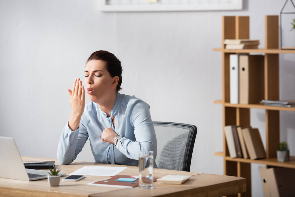 Брюнетка деловая женщина с кулаком на груди кашляет на ладони, сидя на рабочем месте в офисе - Фото, изображение