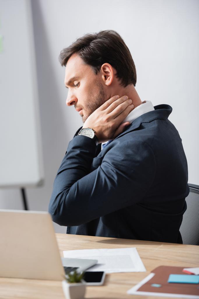 Vue latérale de l'homme d'affaires avec le cou douloureux assis sur le lieu de travail sur fond flou - Photo, image