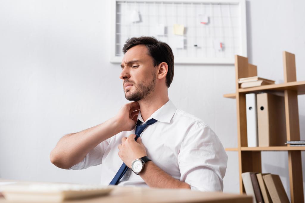 Επιχειρηματίας με επώδυνο λαιμό κάθεται στο χώρο εργασίας με θολή γραφείο σε πρώτο πλάνο - Φωτογραφία, εικόνα