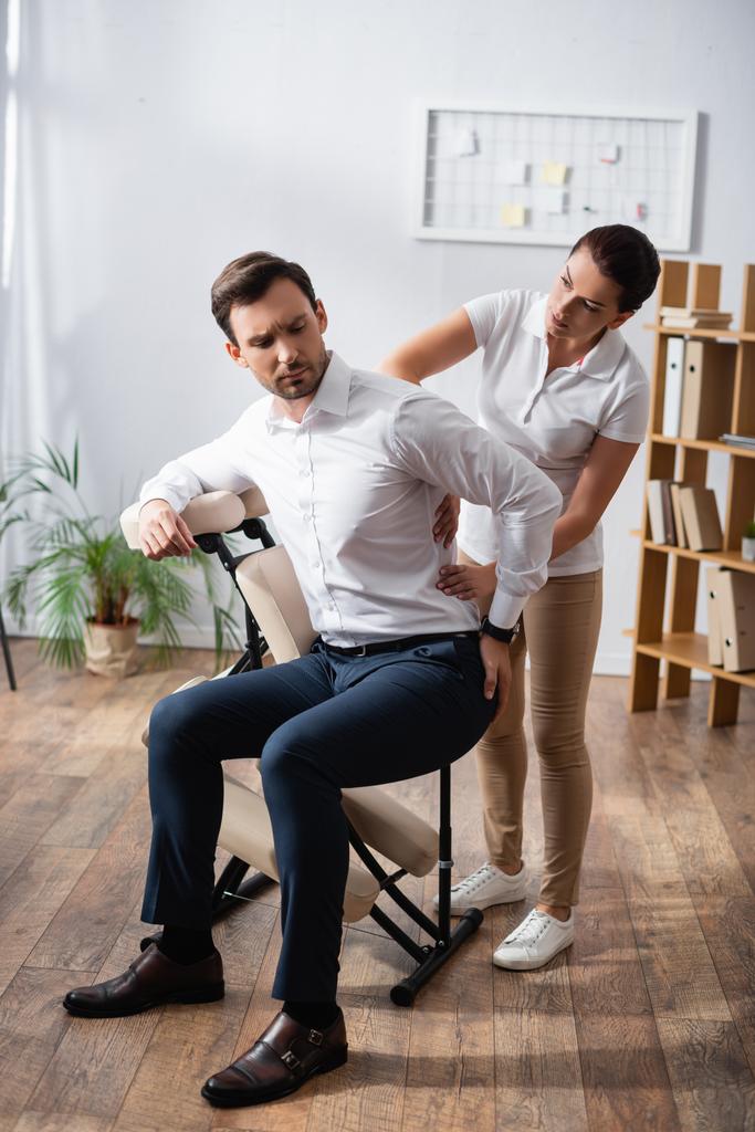 Masajista enfocado masajeando lastimando la espalda del hombre de negocios con la mano en la cadera, sentado en silla de masaje en la oficina - Foto, imagen