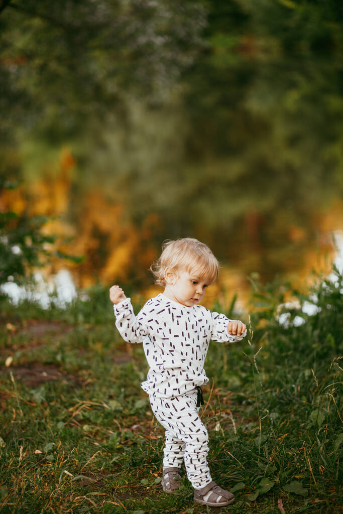 Lindo niño caminando en un hermoso traje blanco contra el fondo de la naturaleza, bebé de alrededor de un año de edad aprendiendo y jugando al aire libre. - Foto, imagen