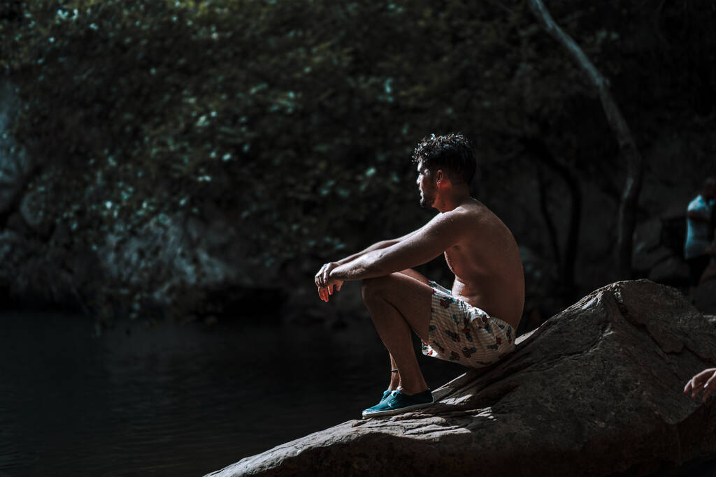 Mladý atraktivní muž v plavkách sedící na skále v lesním jezeře a užívající si přírodní scénu - Fotografie, Obrázek