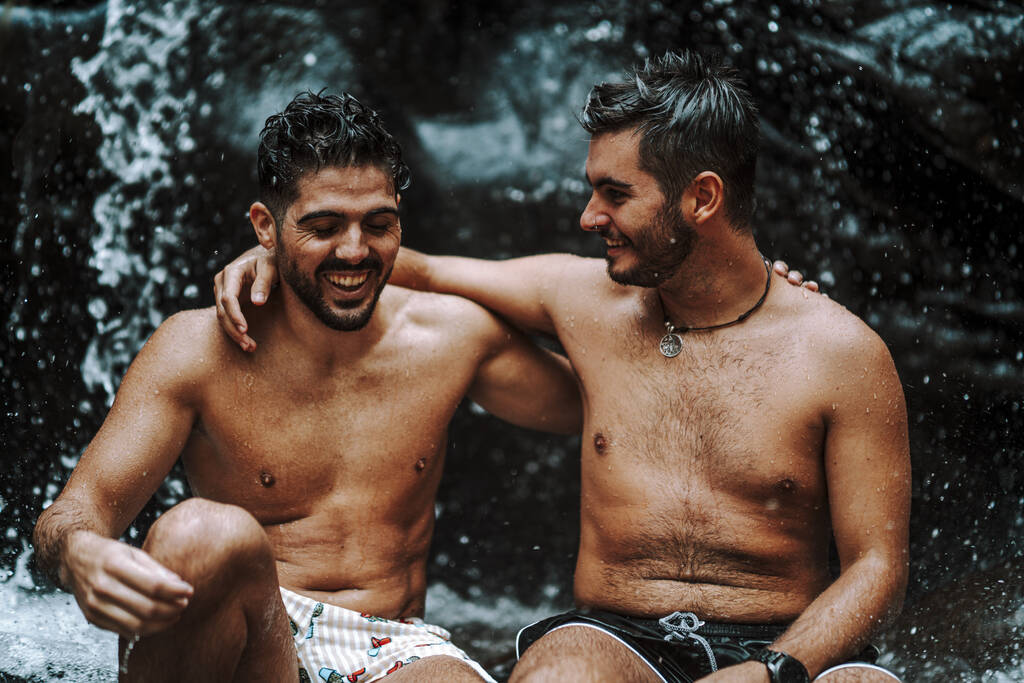 Счастливая гей-пара, сидящая на камне возле прекрасного водопада - Фото, изображение