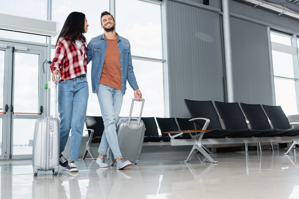 alegre pareja multicultural caminando con equipaje en el aeropuerto  - Foto, Imagen