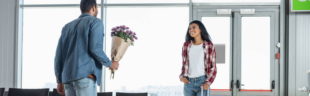 man holding wrapped flowers while meeting happy african american girlfriend in airport, banner - Photo, Image